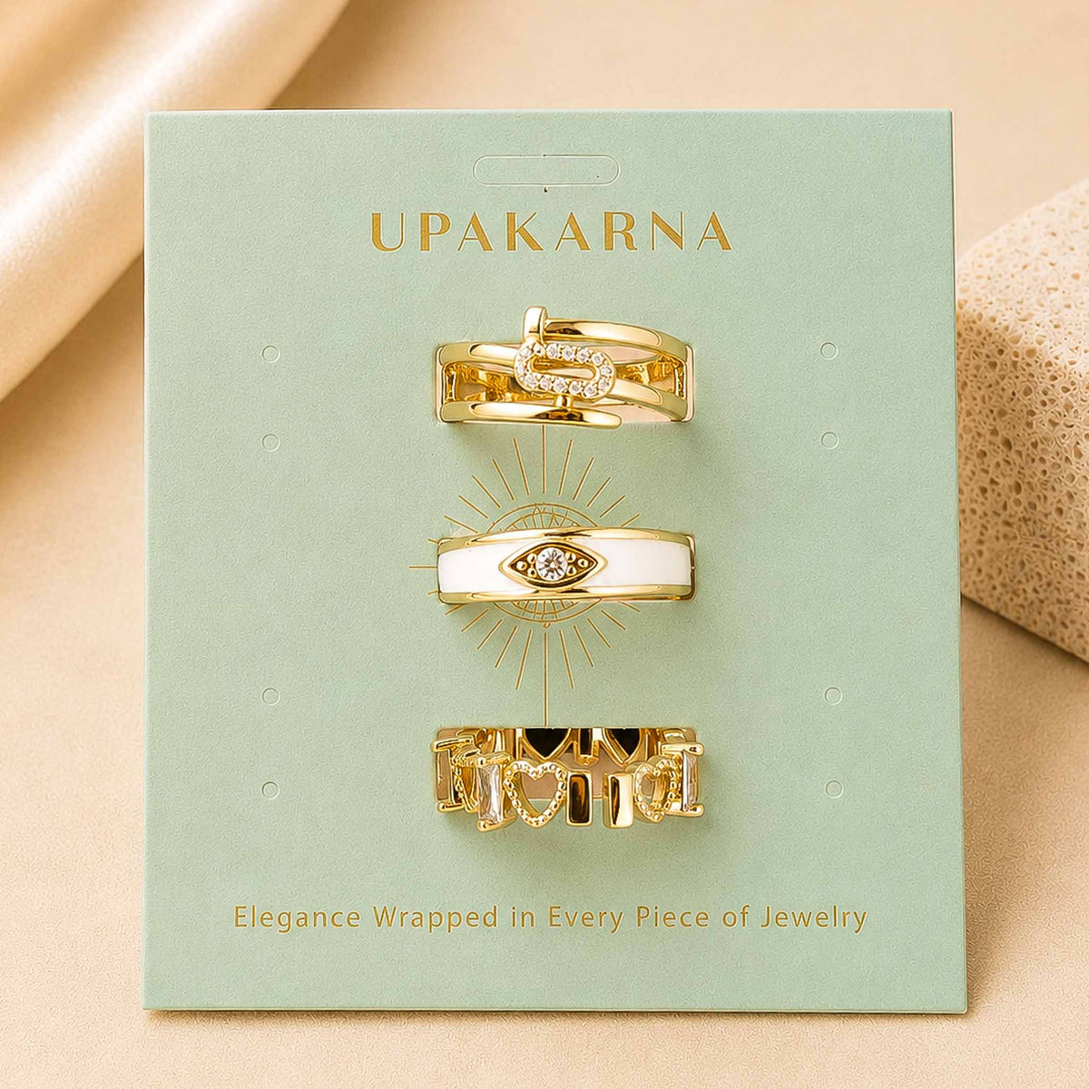 Upakarna jewellery display featuring three gold-toned rings with crystal and evil eye-inspired designs on a sage green branded card.