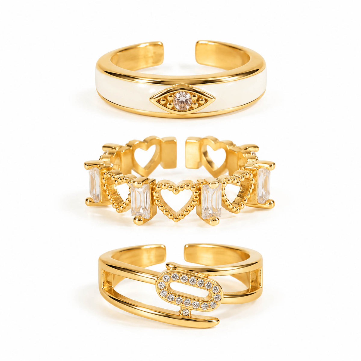 A product photo of three gold-tone adjustable rings featuring a smooth white enamel band with a crystal evil eye symbol, an open band of interconnected filigree hearts and baguette crystals, and a sleek wrap-around band centered with a pave-encrusted oval link.