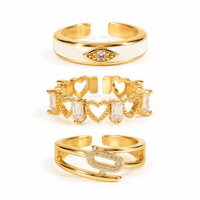 A product photo of three gold-tone adjustable rings featuring a smooth white enamel band with a crystal evil eye symbol, an open band of interconnected filigree hearts and baguette crystals, and a sleek wrap-around band centered with a pave-encrusted oval link.