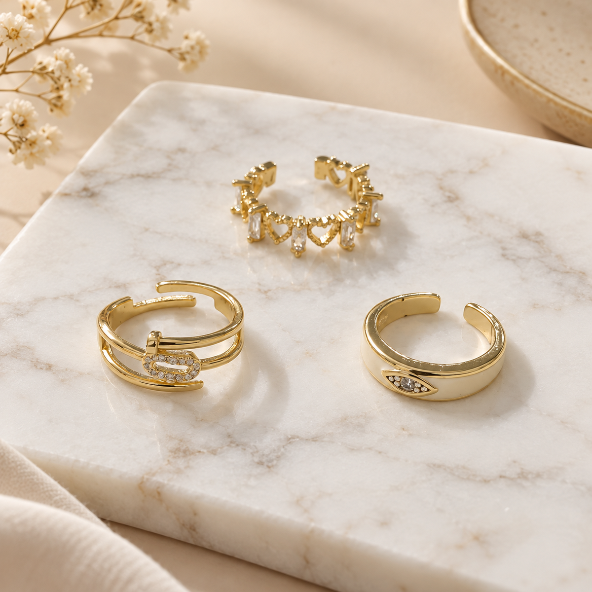 Three gold-tone rings arranged on a white marble surface, featuring polished metal designs with crystal embellishments and delicate heart-shaped details, styled with soft floral accents.