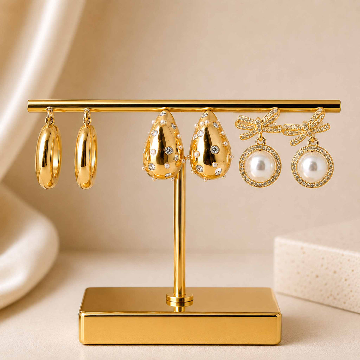 Gold-tone earring stack featuring polished hoops, embellished teardrop earrings, and pearl bow dangler earrings displayed on a gold jewellery stand against a soft beige background.