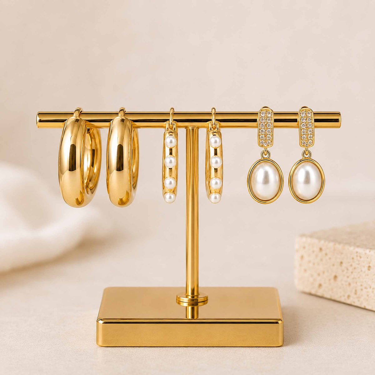 Golden pearl earring stack featuring polished hoops, pearl-lined hoops, and crystal-accented pearl drop earrings, displayed on a gold jewellery stand against a soft beige background.