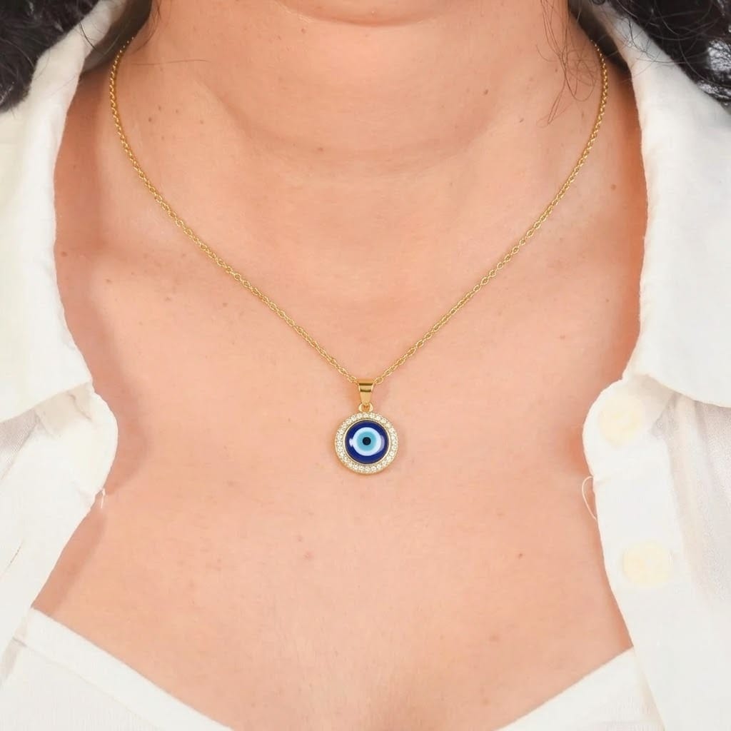 Close-up shot of a person wearing a delicate gold chain necklace with a round Evil Eye pendant. The pendant features a classic blue, light blue, and black eye design surrounded by a halo of clear crystals, resting centrally on the chest over a white button-up collared shirt. The background is a solid, dark gray border.