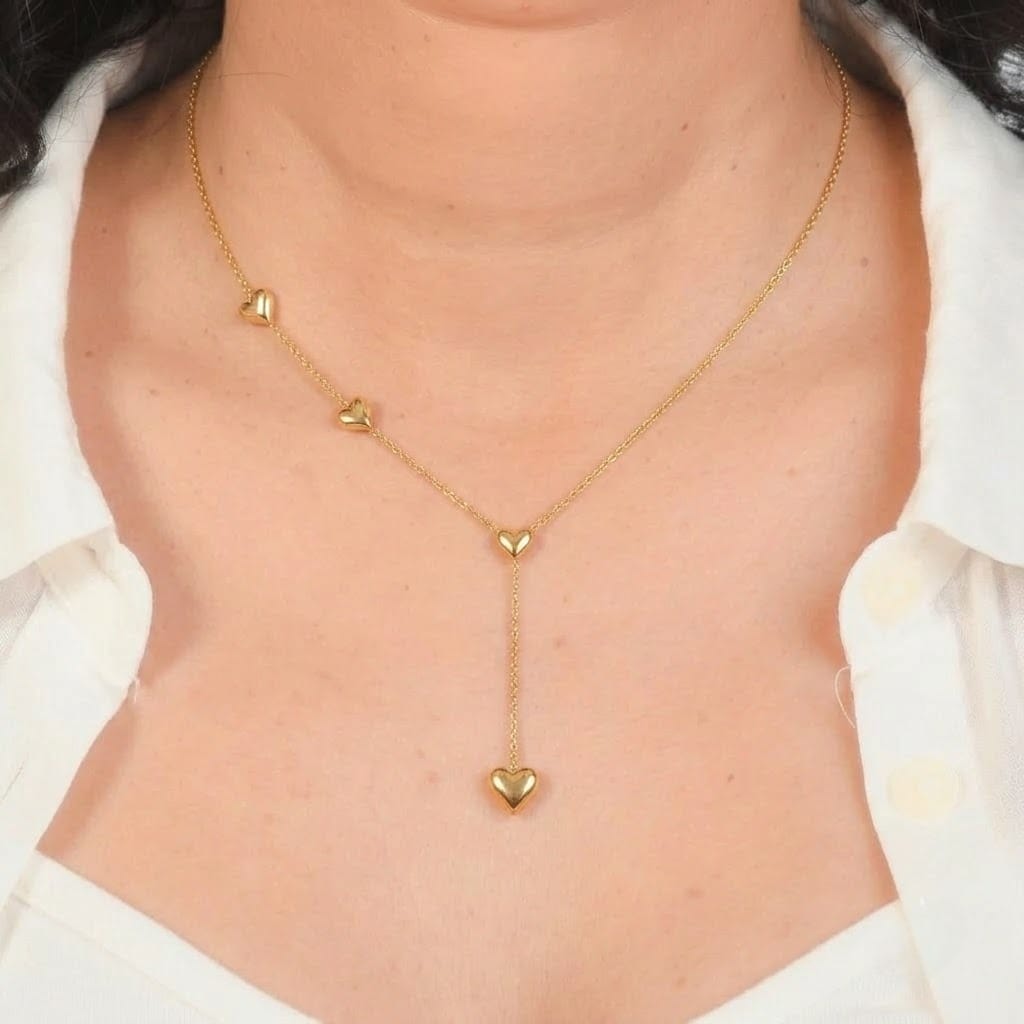 Close-up shot of a model wearing a delicate gold chain lariat necklace featuring small, smooth gold heart charms spaced along the chain and hanging at the drop end. The necklace forms a sleek Y-shape on the chest, framed by an open white collared shirt against a dark gray background frame.
