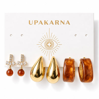 Three pairs of gold-tone statement earrings displayed on an Upakarna jewellery card, featuring crystal bow earrings with brown pearl drops, oversized polished teardrop earrings, and glossy amber resin hoop earrings, arranged on a clean white background with a premium polished finish.