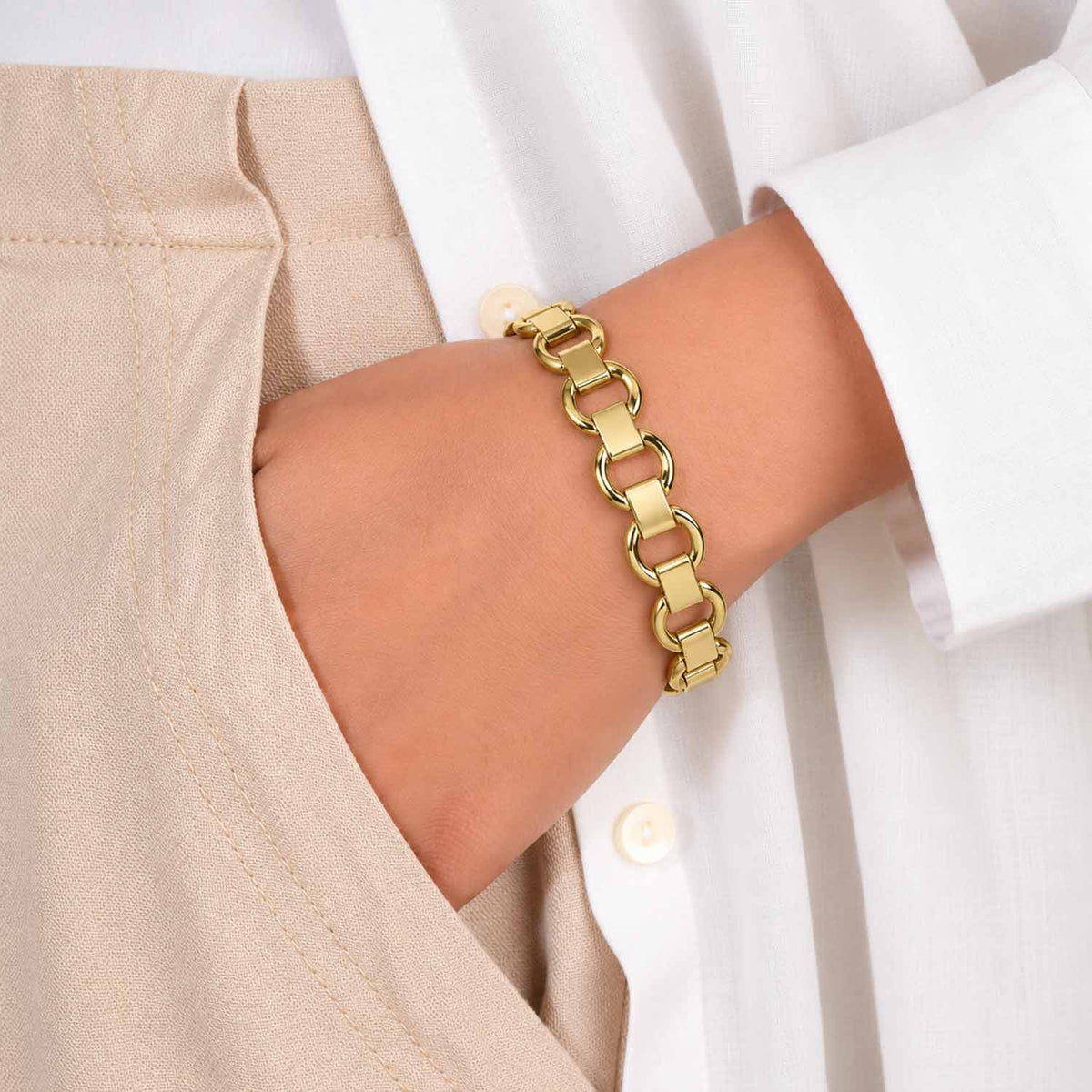 Gold cuff bangle featuring an interlocking rectangular and circular link design, worn on the wrist with a neutral beige and white outfit.