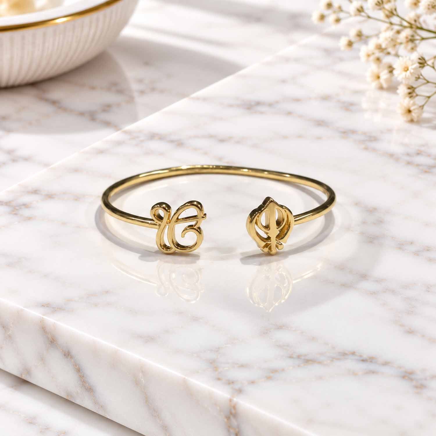 Gold open cuff bangle featuring an Ik Onkar symbol and Khanda motif, displayed on a marble surface.