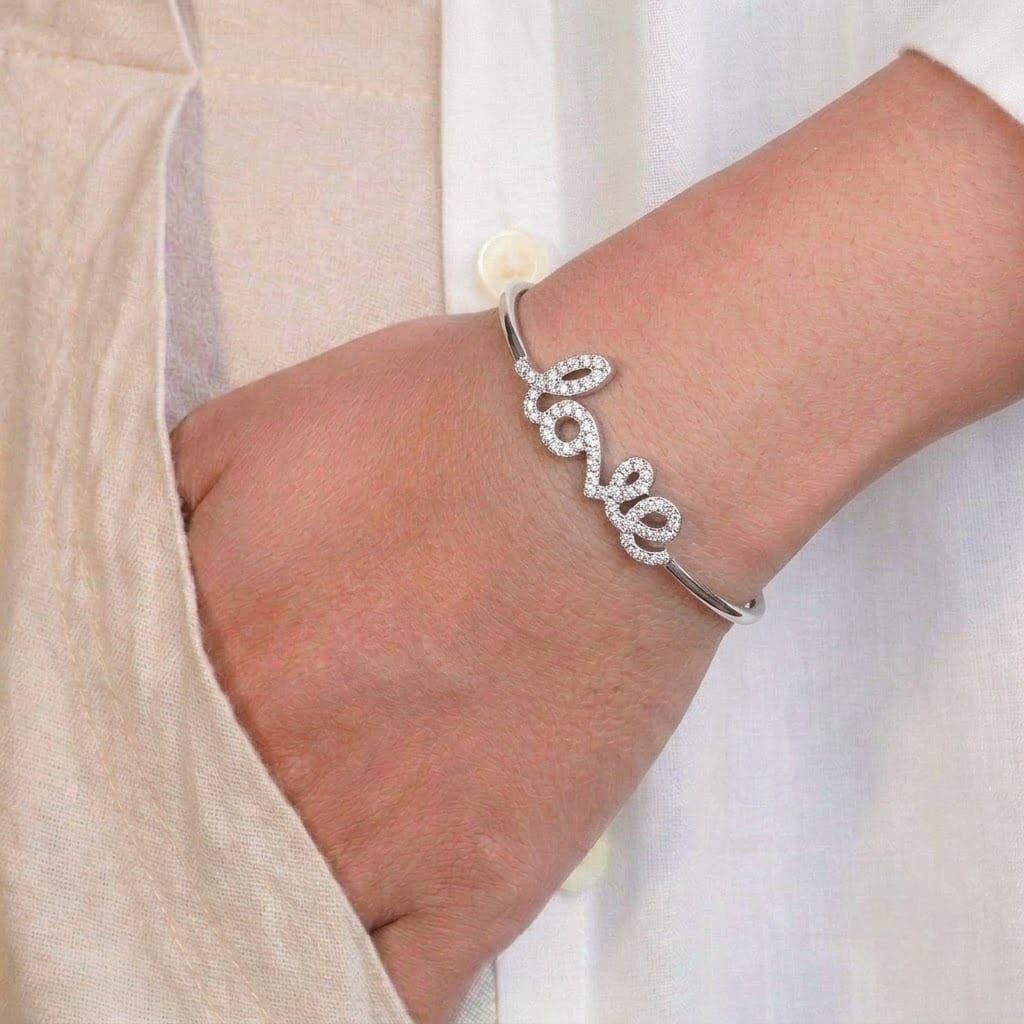 A close-up of a person’s wrist wearing a sleek silver-toned bangle featuring the word “love” in a cursive script, embellished with small sparkling stones. The minimalist bracelet is styled against a soft beige outfit, creating an elegant and delicate look.