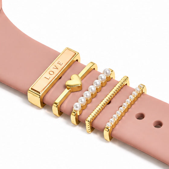 Zoomed-in view of gold-tone watch charms featuring a LOVE charm, heart charm, pearl accents, textured chain detail, and pearl-lined design on a blush pink watch strap against a clean white background.