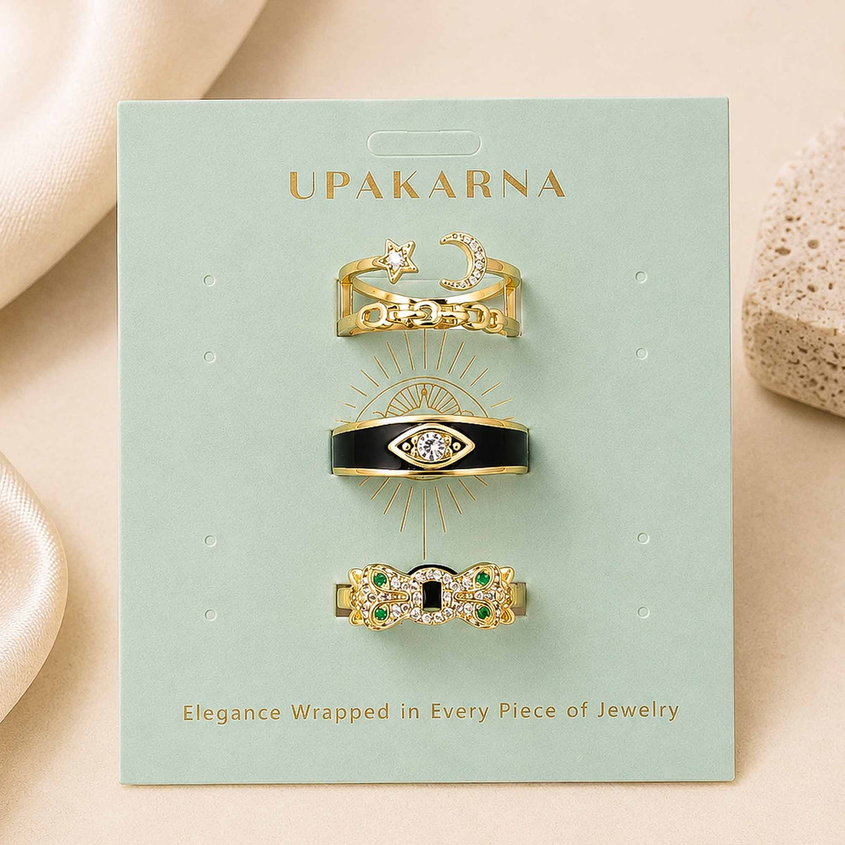 Upakarna jewellery display featuring three gold-toned statement rings with star and moon, black evil eye, and green gemstone bow designs on a sage green branded card.