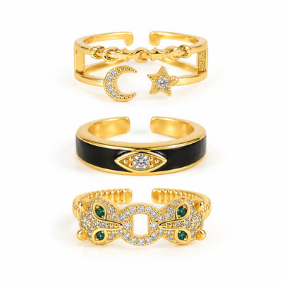 A product photo of three gold-tone adjustable rings featuring an open band with dangling celestial charms, a smooth band with black enamel and a crystal-set evil eye motif, and a textured band encrusted with crystals and green gemstone accents.