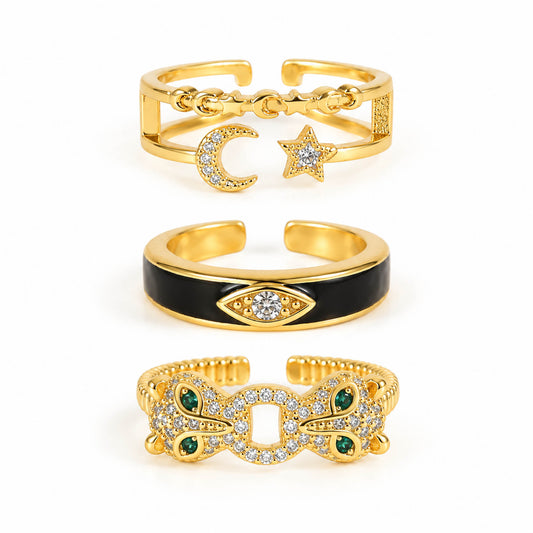 A product photo of three gold-tone adjustable rings featuring an open band with dangling celestial charms, a smooth band with black enamel and a crystal-set evil eye motif, and a textured band encrusted with crystals and green gemstone accents.