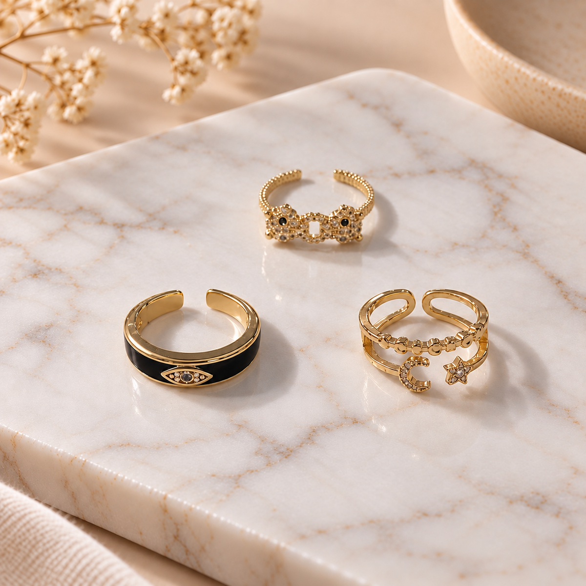 Three gold-tone rings arranged on a white marble surface, featuring polished metal designs with crystal embellishments and delicate decorative details, styled with soft floral accents.