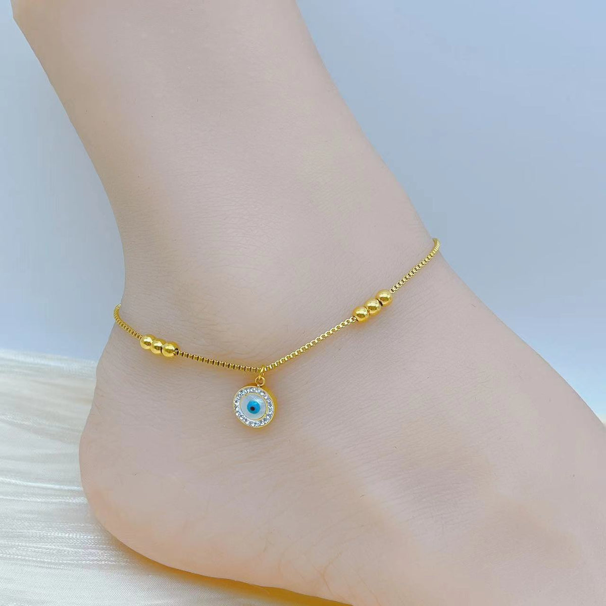Delicate gold-tone anklet featuring a round blue evil eye pendant surrounded by sparkling clear stones, with small polished gold beads along a fine chain, styled on an ankle against a soft blue and white background.