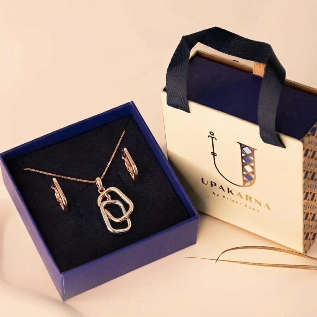 A dual-tone jewelry set featuring a delicate rose gold chain necklace with interlocking abstract link pendants in rose gold and silver. The set includes matching small hoop earrings, displayed inside an open navy blue box. Beside it is an elegant Upakarna branded gift bag in cream and navy with ribbon handles, placed on a soft beige background for a premium presentation.