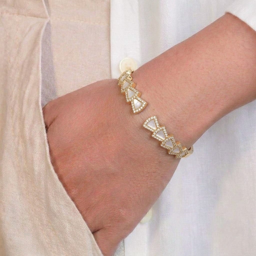 A close-up of a person’s wrist wearing a delicate gold-toned bangle featuring a series of small triangular motifs, each outlined with tiny sparkling stones. The geometric bracelet is styled against a soft beige outfit, giving a modern and elegant appearance.