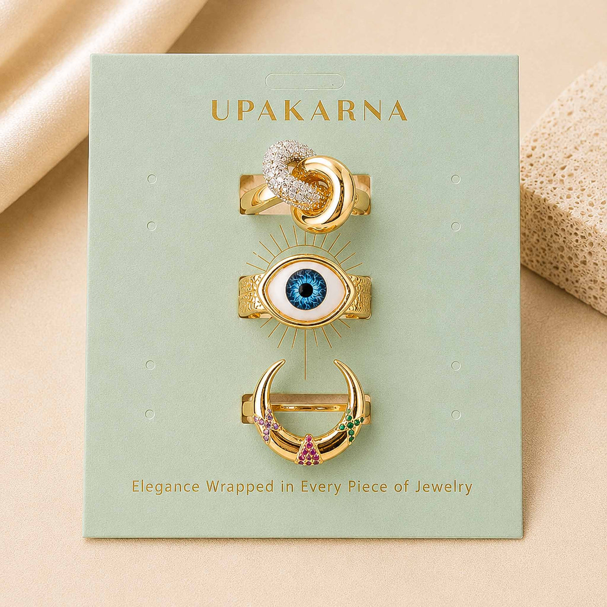 Gold-toned celestial and evil eye rings displayed on a mint green Upakarna jewellery card, featuring moon, crystal, blue eye, and colourful gemstone designs.