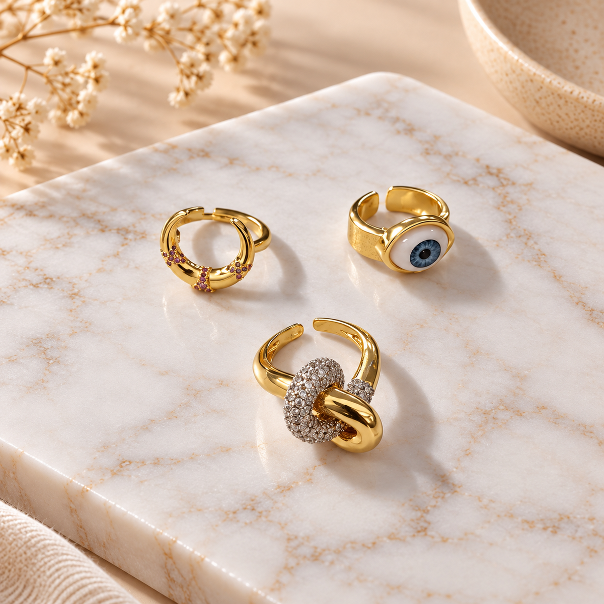 Three gold tone rings arranged on a white marble surface with soft natural lighting and delicate floral accents in the background.