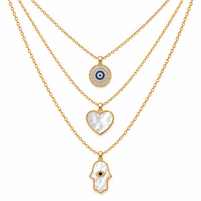 A product photo of a layered gold-tone necklace set featuring a circular evil eye pendant surrounded by rhinestones, a heart-shaped white mother-of-pearl pendant, and a white mother-of-pearl Hamsa hand pendant with a crystal-accented center.   