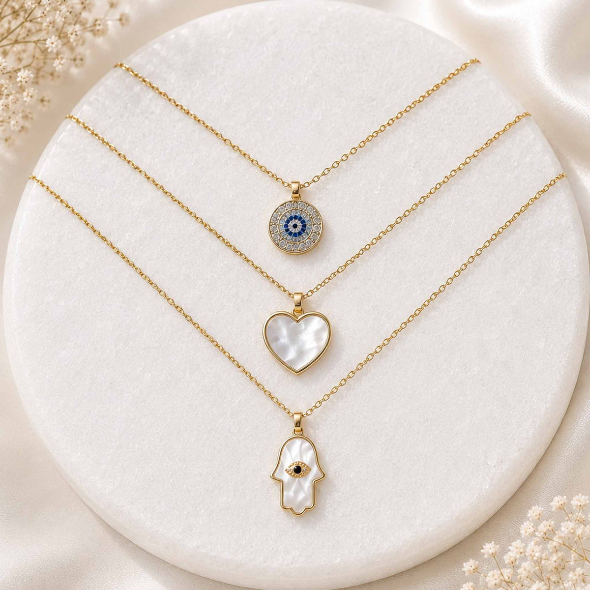 Gold layered necklace set with evil eye, mother-of-pearl heart, and Hamsa hand pendants displayed on a white circular jewellery stand.