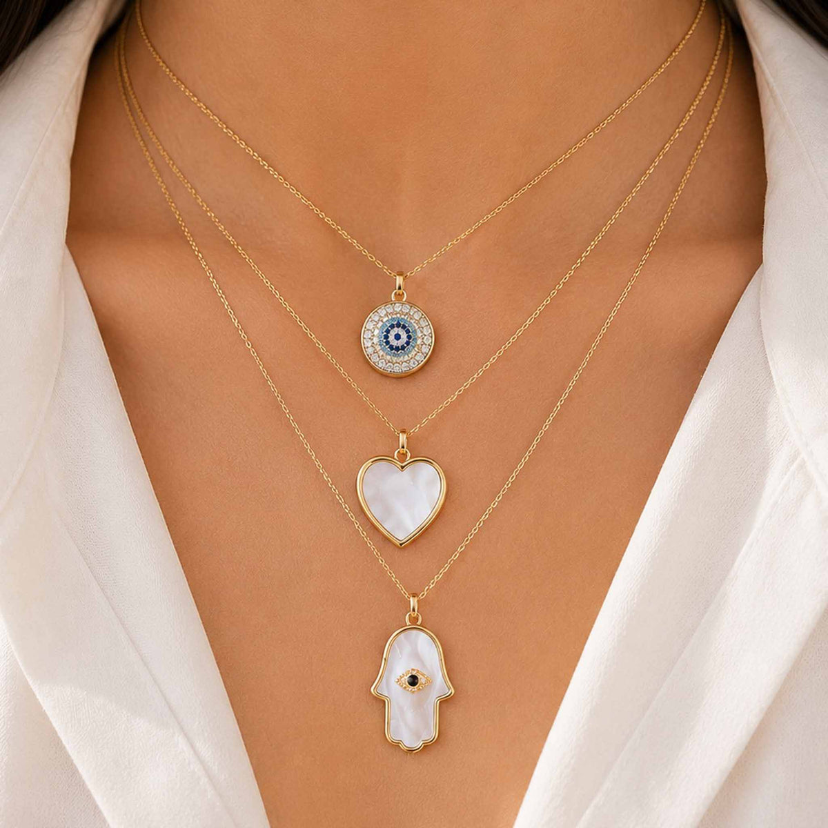 Gold layered necklace set with evil eye, heart, and Hamsa hand pendants worn with a white outfit.