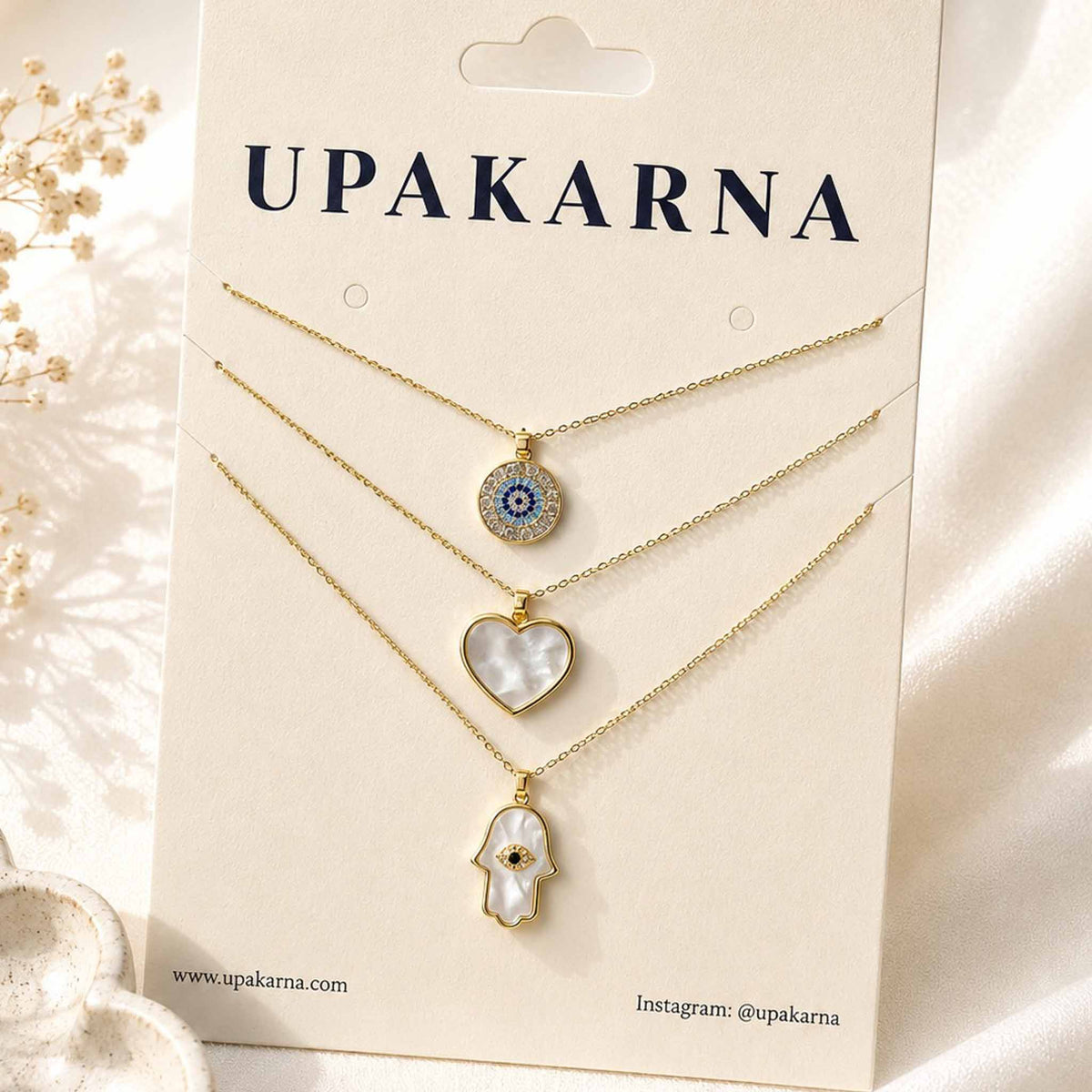 Gold layered necklace set featuring evil eye, mother-of-pearl heart, and Hamsa hand pendants displayed on an Upakarna jewellery card.