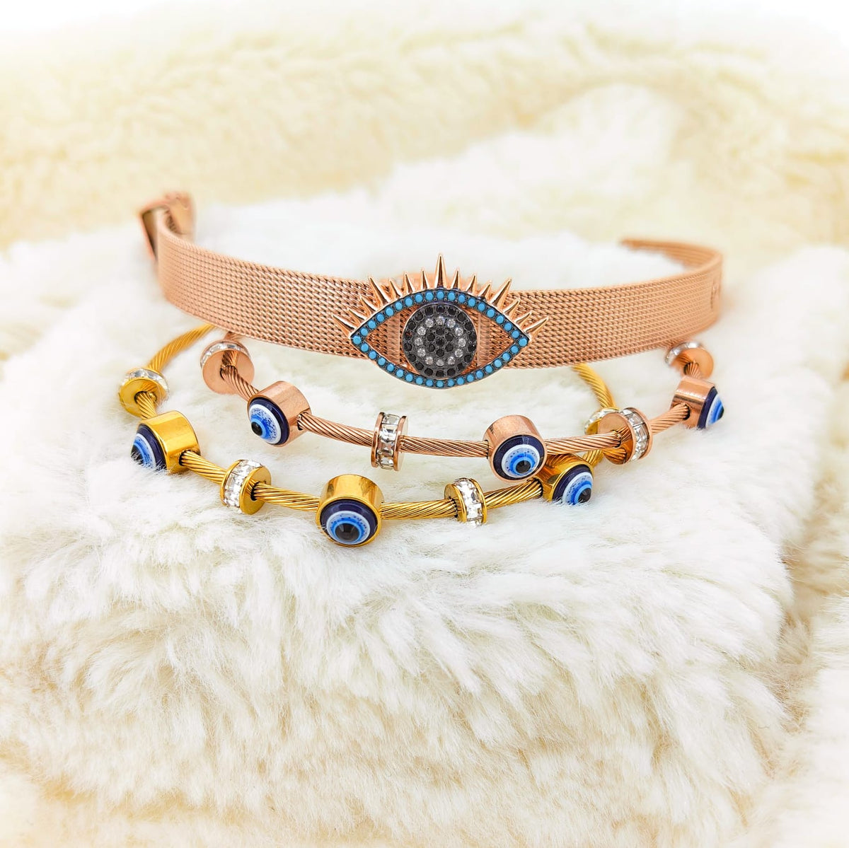 A studio product photograph of a curated stack of mixed-metal charm bracelets resting on a fluffy white background. The set features a rose gold-toned mesh-band bracelet centered with an elaborate pavé-set evil eye charm flanked by turquoise accents, layered over two cable-wire bracelets in gold and rose-gold tones accented with dark blue and white evil eye beads and sparkling crystal station spacers.   