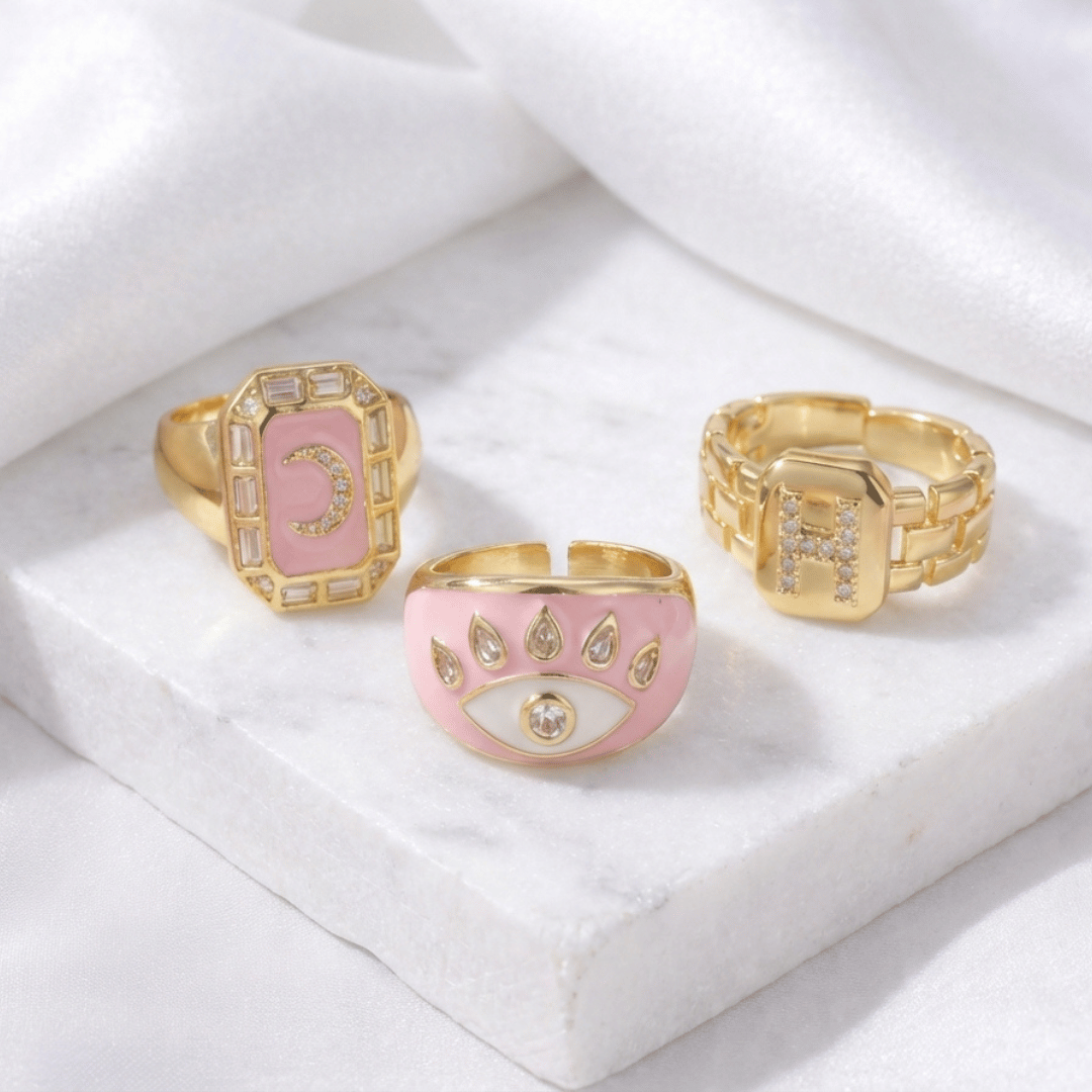 A stack of three adjustable gold rings featuring a pastel evil eye, a celestial moon motif, and a personalized initial charm.