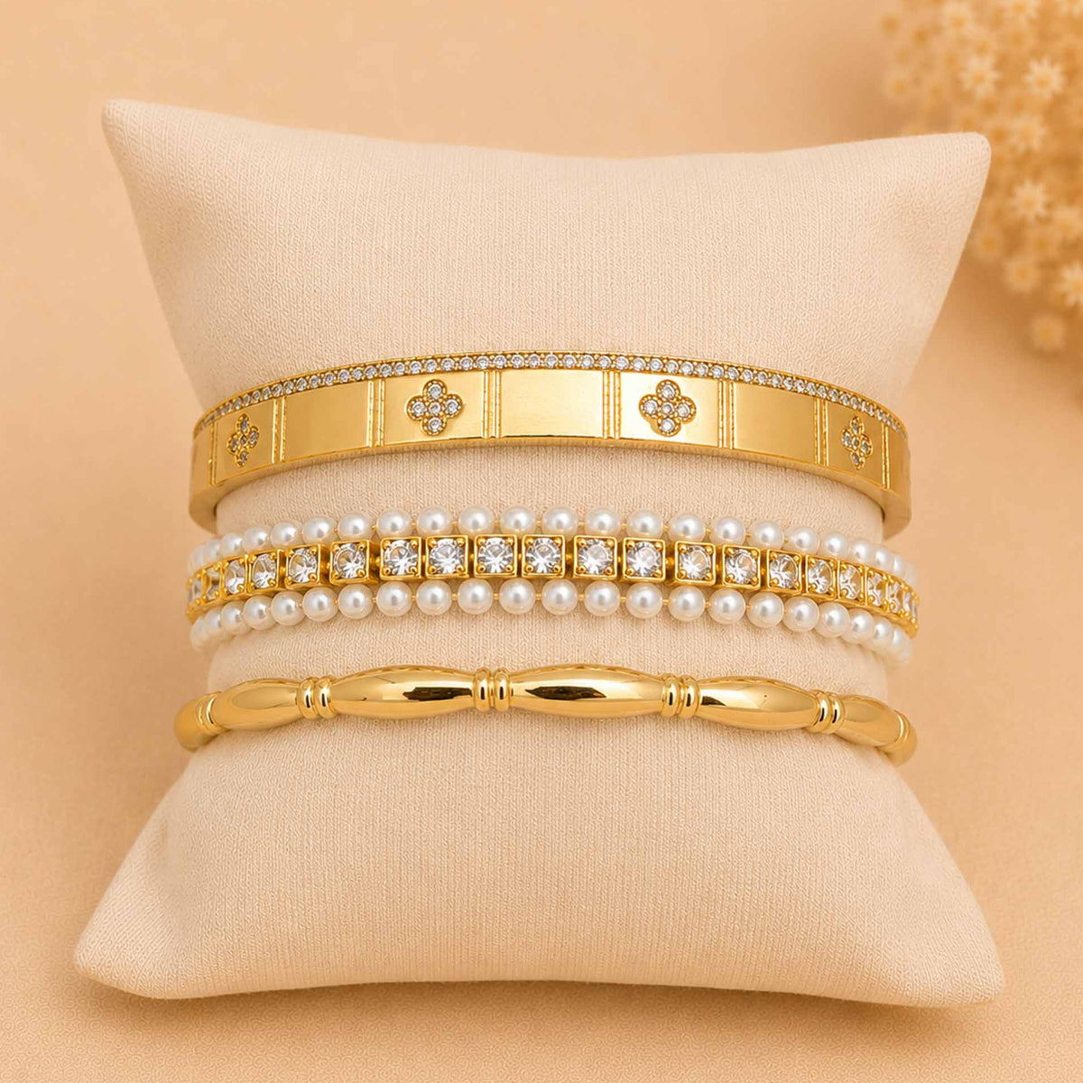 Gold bangle stack displayed on a cream cushion, featuring a wide embellished cuff, pearl and crystal bracelet, and polished gold bangle.
