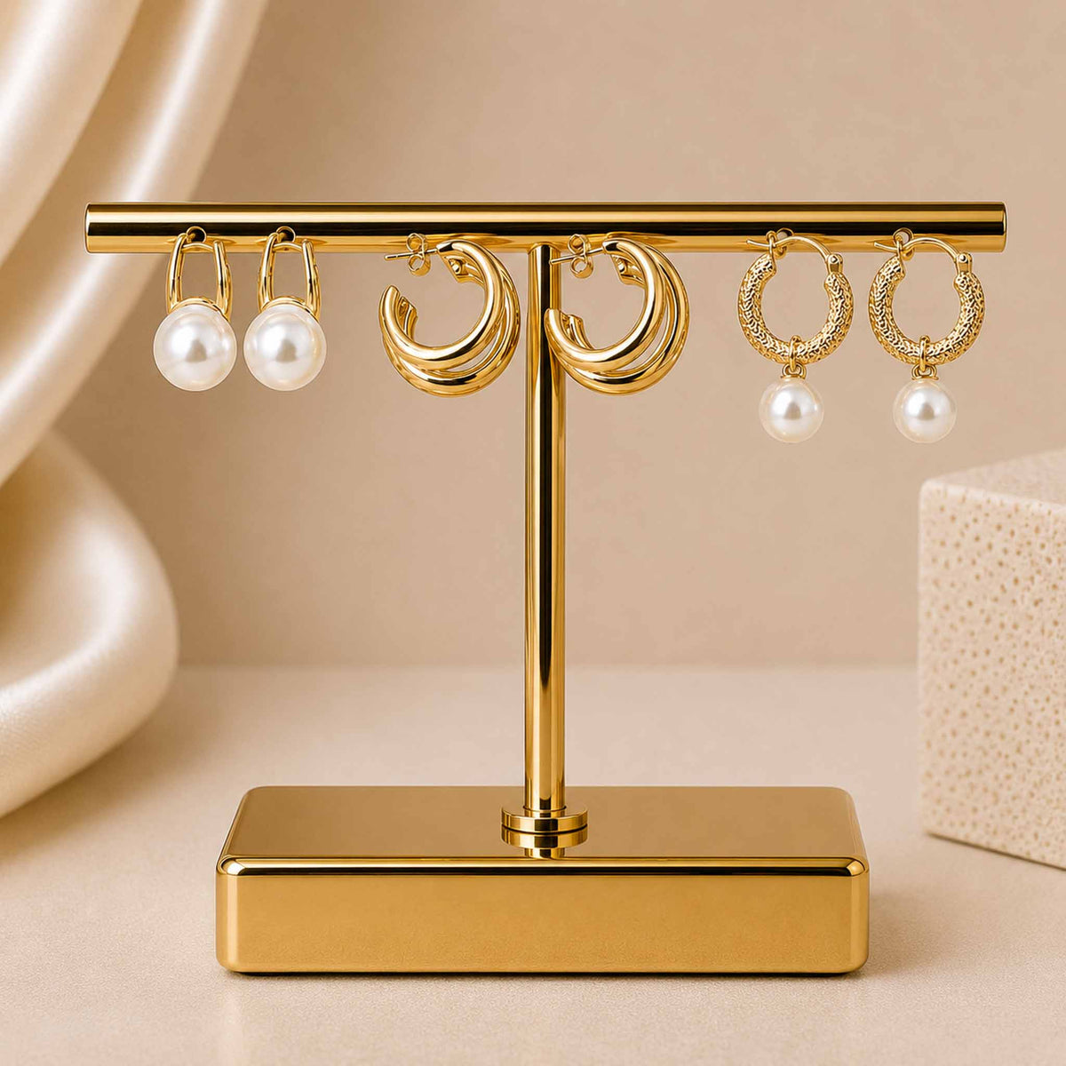 Gold-tone earring stack featuring pearl stud earrings, layered gold hoop earrings, and textured pearl drop hoop earrings displayed on a gold jewellery stand against a soft beige background.
