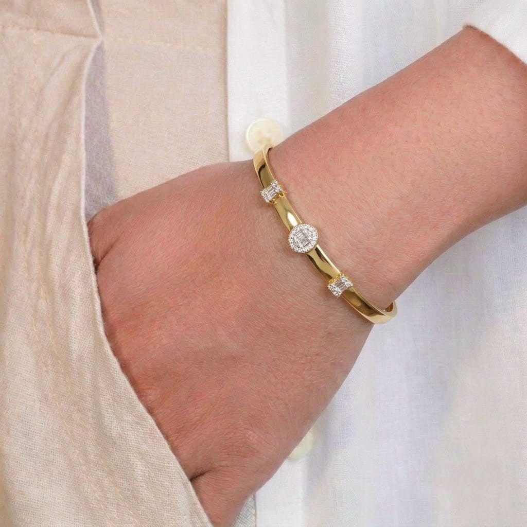 An anti-tarnish yellow gold cuff bracelet styled on a model's wrist resting against a neutral linen shirt. The sleek, highly polished gold band features three distinct crystal accent stations: a central oval halo motif set with baguette and round cubic zirconia stones, flanked on either side by rectangular baguette-cut cluster stations for a balanced, sophisticated sparkle.