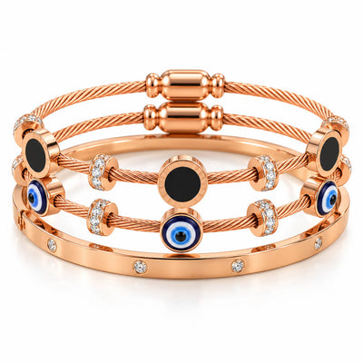 A studio shot of a curated luxury bracelet stack featuring multiple rose gold cable wire strands adorned with black circular enamel stations, blue-and-white evil eye charms, and sparkling rhinestone spacers, paired with a matching gem-set bangle.