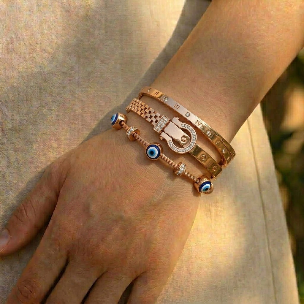 Luxury rose gold anti-tarnish bracelet stack featuring a Roman numeral bangle, crystal-studded buckle bracelet, textured chain bangle, and a blue evil eye cable bracelet, elegantly worn on the wrist in natural sunlight.