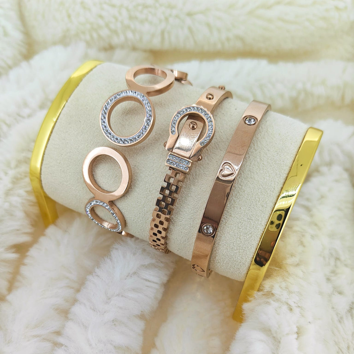 Rose gold anti-tarnish bracelet stack featuring an elegant buckle-style crystal bracelet, a sleek heart-detail bangle with sparkling stones, and a statement oval-link bracelet with crystal accents. Displayed on a cream jewellery cushion with polished gold-tone stands against a soft ivory faux-fur background for a luxurious editorial finish.