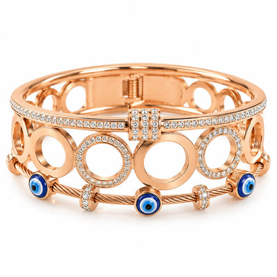 A studio shot of a curated stack of luxury rose gold bracelets, showcasing an openwork circular link pattern, a shimmering rhinestone-paved band, and a twisted cable bracelet accented with enamel evil eye beads.