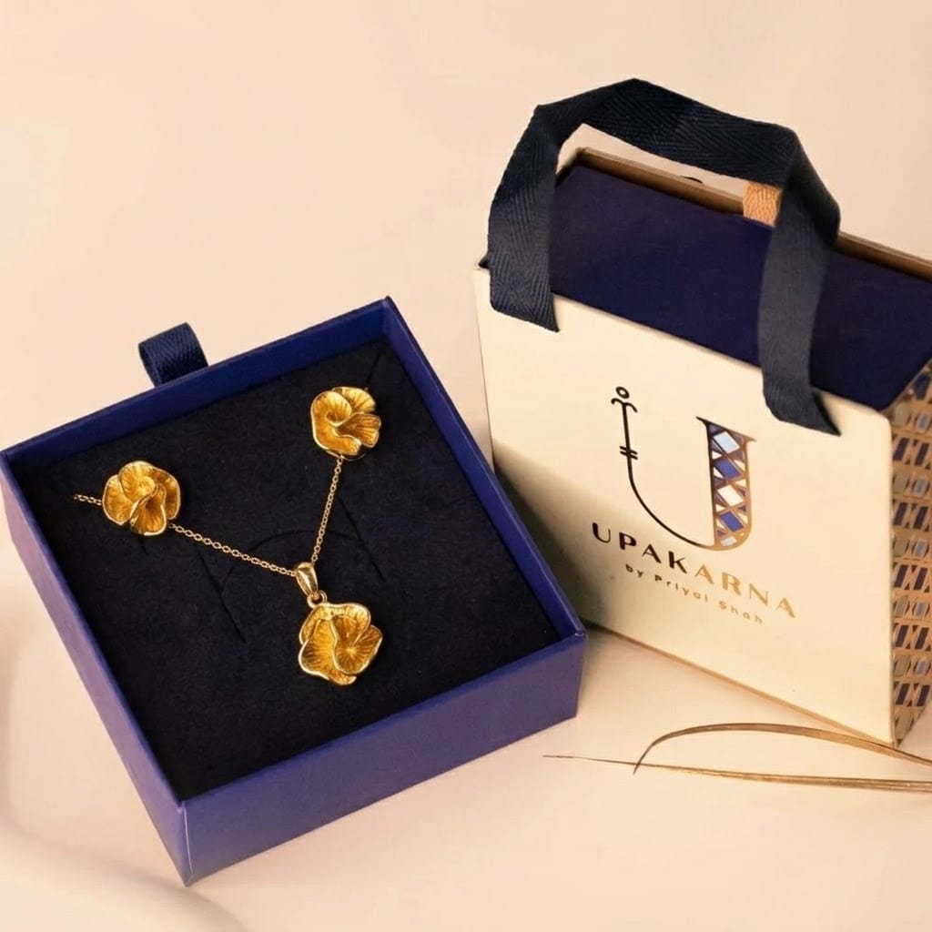 A gold-tone jewelry set with a textured floral design, featuring a delicate chain necklace and matching stud earrings, displayed inside an open navy blue box. The set is accompanied by a branded Upakarna gift bag in cream and navy with ribbon handles, placed on a soft beige background, creating a premium and elegant presentation.