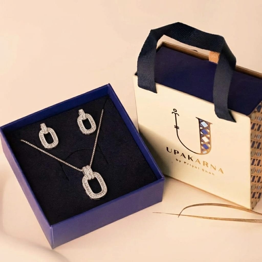 A silver jewelry set presented inside a dark blue display box next to a cream "UPAKARNA by Priyal Shah" gift bag. Arranged on the navy velvet interior, the set features a box-chain necklace with an open oval pendant bordered in baguette-cut cubic zirconia and attached to a double-row pavé bail, accompanied by a pair of matching oval drop earrings.