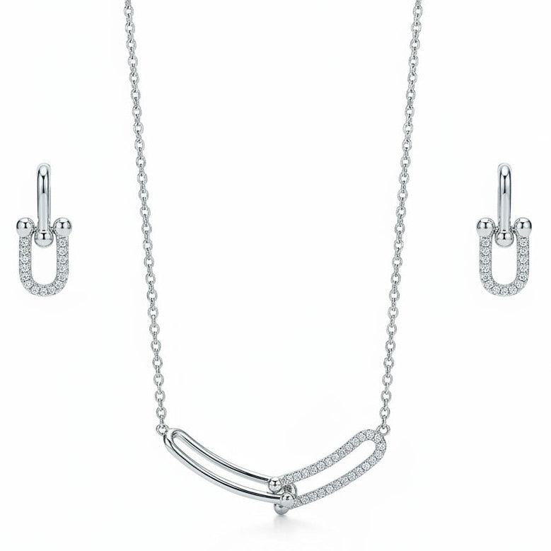 A silver jewelry set isolated on a plain white background, featuring a delicate fine-chain necklace with a curved central pendant of two interlocking U-shaped links—one smooth and polished, the other encrusted with micro-pavé cubic zirconia stones—paired with matching U-shaped drop earrings with pavé crystal accents.