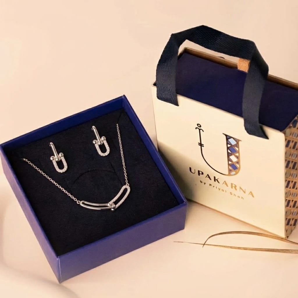 A silver jewelry set presented inside a dark blue display box next to a cream "UPAKARNA by Priyal Shah" gift bag. Set on the navy interior tray, the collection includes a fine-chain necklace featuring an interlocking U-shaped link pendant with micro-pavé cubic zirconia accents, alongside a pair of matching U-link drop earrings.