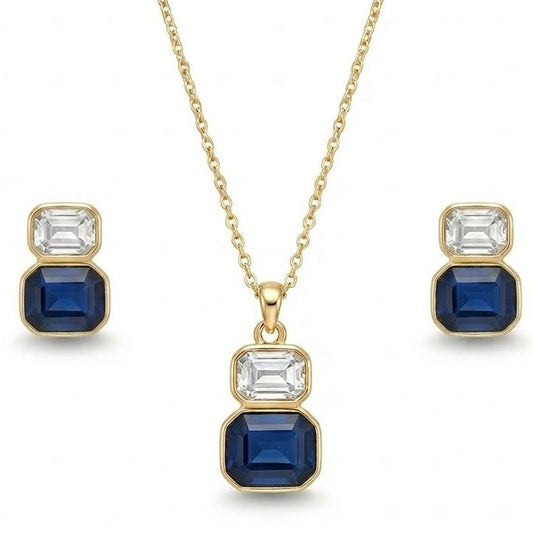 A gold jewelry set isolated on a plain white background, featuring a fine-chain necklace with a two-stone drop pendant consisting of a clear, emerald-cut crystal stacked atop a larger sapphire-blue emerald-cut gemstone, both set in smooth gold bezels. The set is paired with matching stud earrings that replicate the same stacked clear and blue stone design.