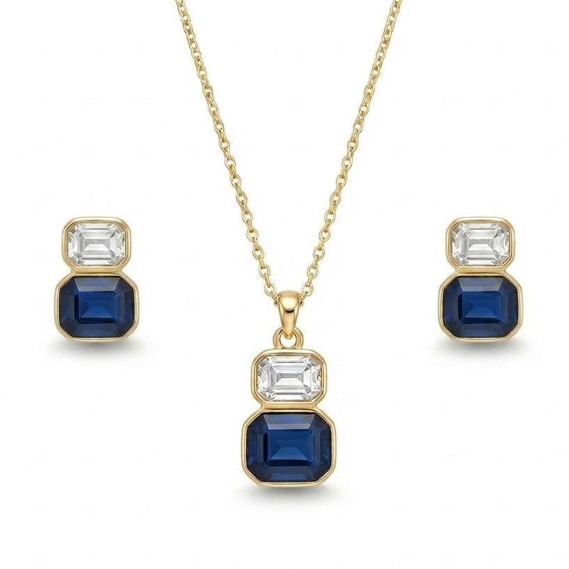 A gold jewelry set isolated on a plain white background, featuring a fine-chain necklace with a two-stone drop pendant consisting of a clear, emerald-cut crystal stacked atop a larger sapphire-blue emerald-cut gemstone, both set in smooth gold bezels. The set is paired with matching stud earrings that replicate the same stacked clear and blue stone design.
