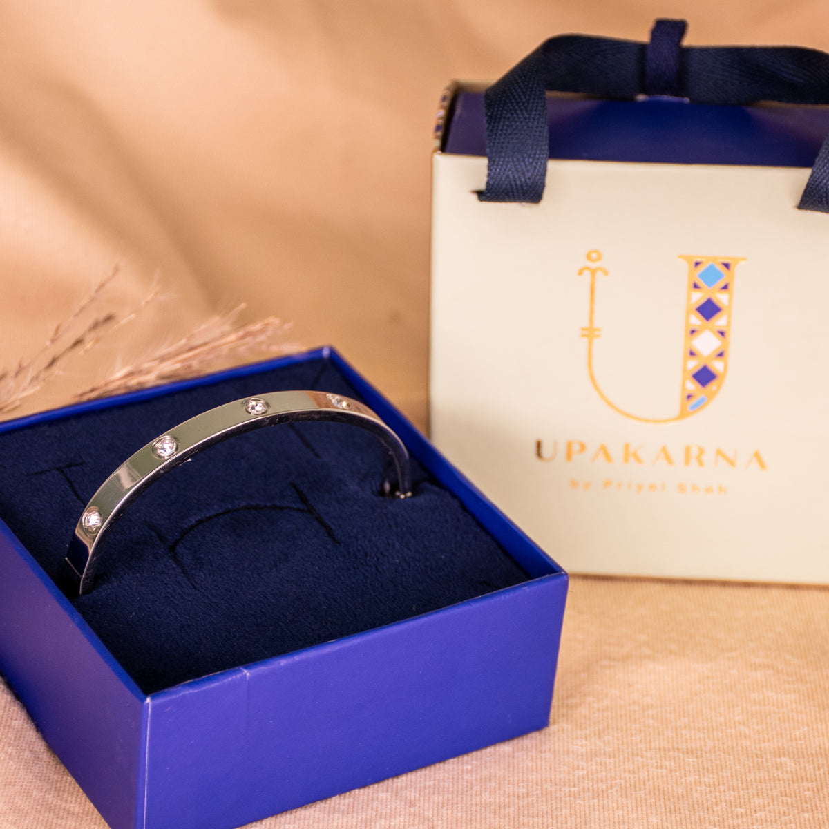 Silver anti-tarnish bangle featuring a sleek polished finish and sparkling crystal accents, displayed in an open navy blue Upakarna jewellery box with a cream gift bag featuring the brand’s gold logo. Styled against a warm beige background with delicate dried botanicals and soft golden lighting for a premium gifting aesthetic.