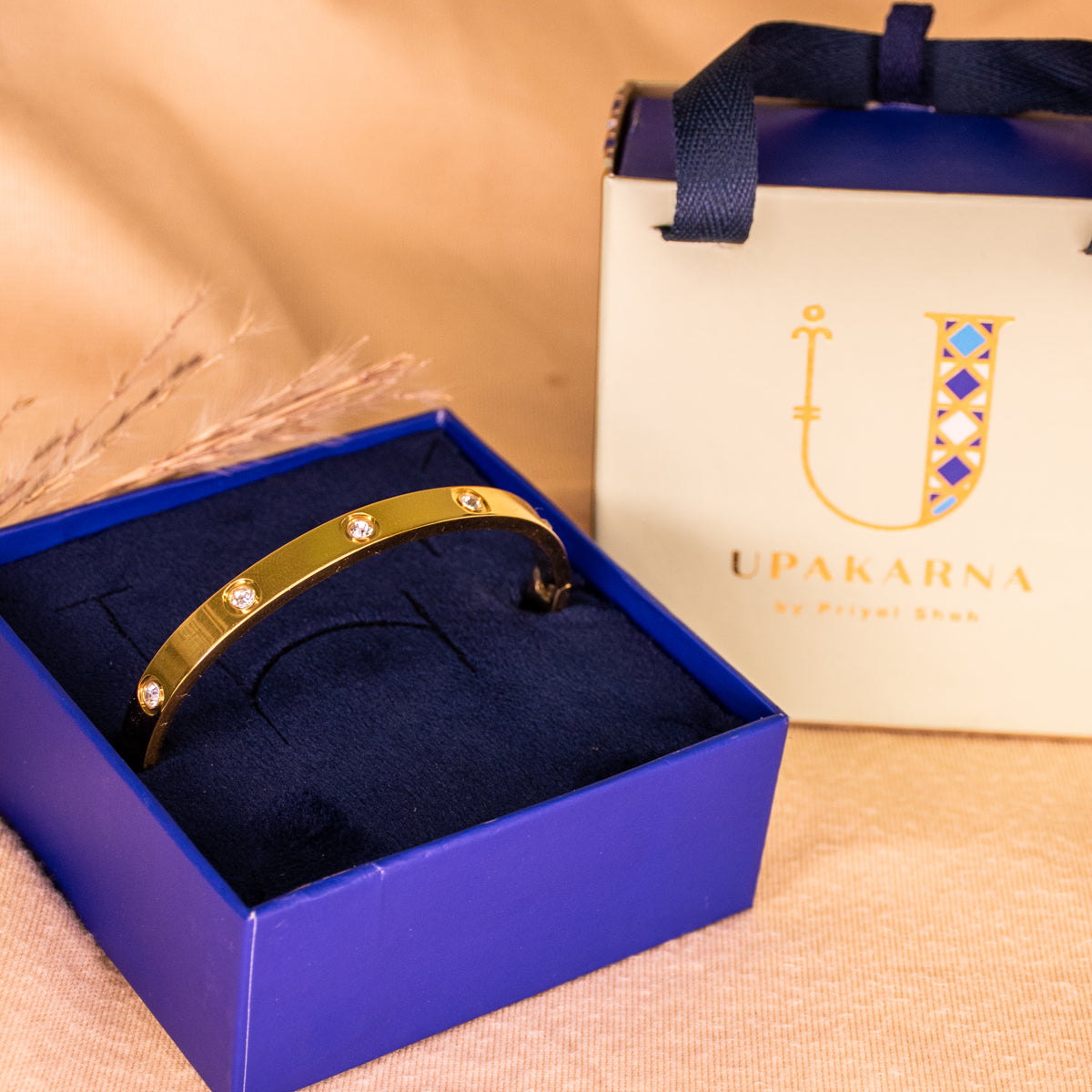 Gold anti-tarnish bangle featuring a sleek polished finish and sparkling crystal accents, presented in an open navy blue Upakarna jewellery box with a cream branded gift bag. Styled against a warm beige background with delicate dried botanicals and soft golden lighting for a premium, elegant gifting aesthetic.