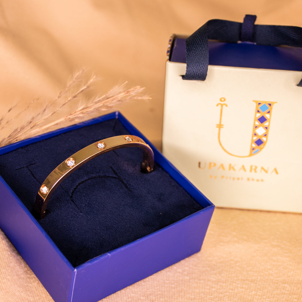 RoseGold anti-tarnish bangle featuring a sleek polished finish and sparkling crystal accents, elegantly presented in an open navy blue Upakarna jewellery box with a cream branded gift bag. Styled against a warm beige background with delicate dried botanicals and soft golden lighting for a premium luxury gifting aesthetic.