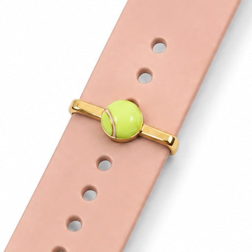 Close-up view of a lime green tennis ball watch charm with gold-tone detailing, attached to a blush pink watch strap on a clean white background.