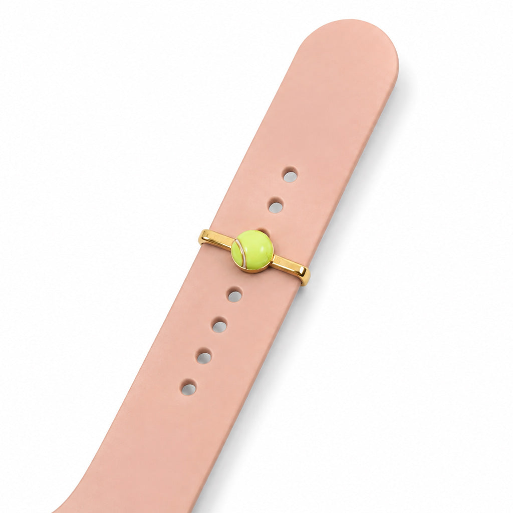Zoomed-out view of a lime green tennis ball watch charm with gold-tone detailing, attached to a blush pink watch strap on a clean white background.