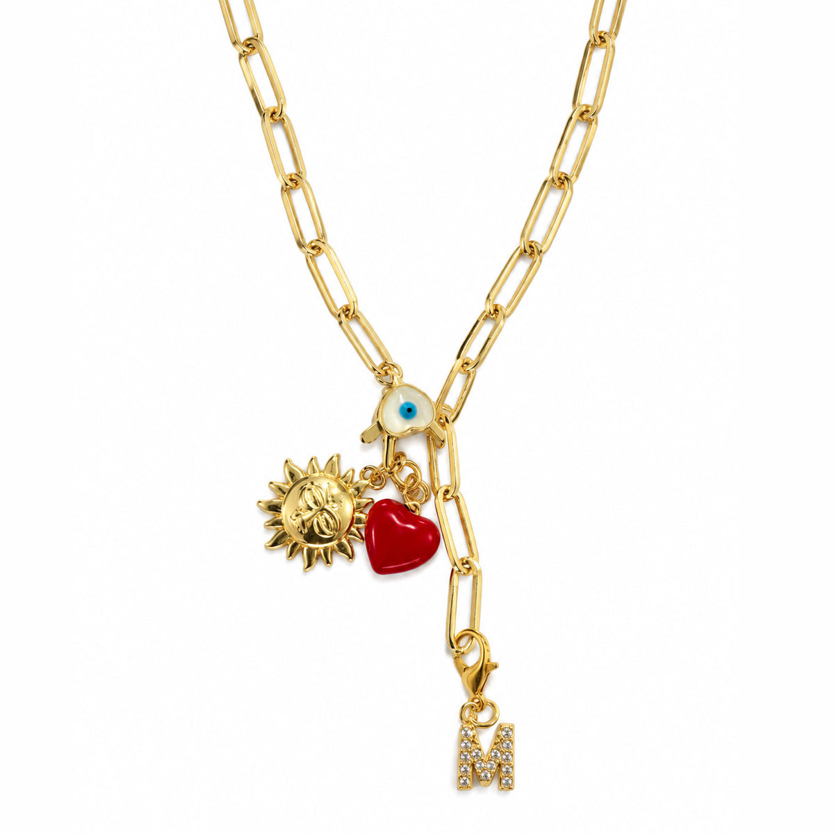 Gold-plated paperclip chain necklace with a blue Evil Eye centerpiece, dangling sun and red heart charms, and a sparkling “M” initial charm, styled on a clean white background.