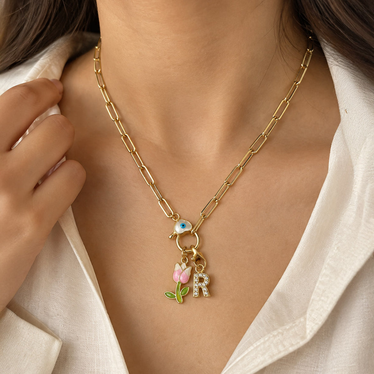 Gold-plated paperclip chain necklace worn on a model, featuring a blue Evil Eye centerpiece with dangling pink tulip and sparkling “R” initial charms.

