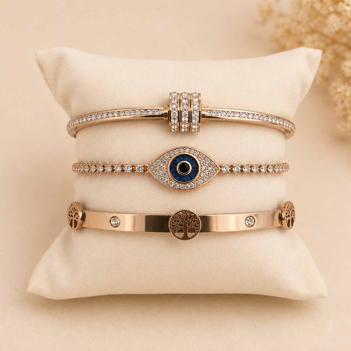 Rosegold bangle stack featuring a sparkling twisted bangle, Evil Eye tennis bracelet, and Tree of Life bangle displayed on a cream cushion.