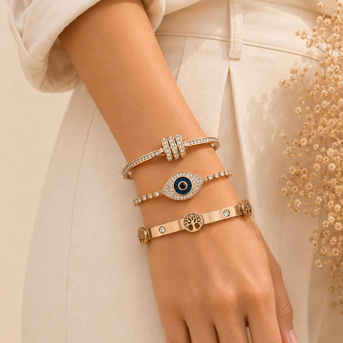 Rosegold bangle stack worn on a wrist, featuring a sparkling twisted bangle, Evil Eye tennis bracelet, and Tree of Life bangle with a cream outfit.